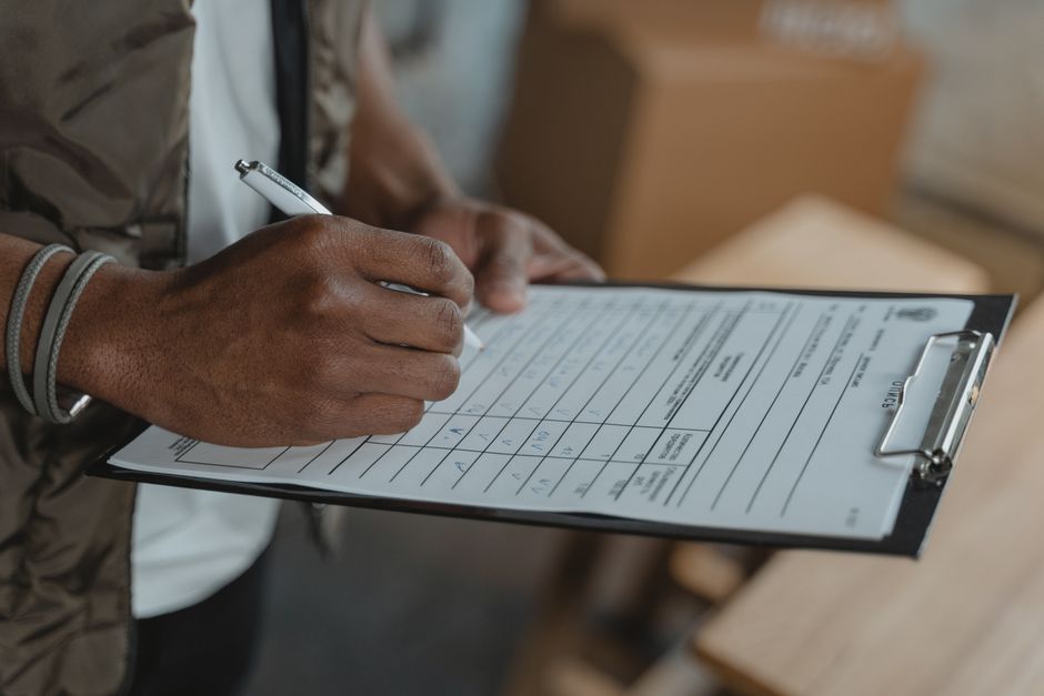 Close-up of a person's hands holding a clipboard with a detailed moving checklist, using a pen to fill out or review the form. The individual is wearing a brown jacket and a gray bracelet on their lef