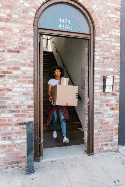 A woman with curly hair, dressed in casual clothing including a white t-shirt, blue jeans, and white sneakers, is descending a staircase at the entrance of a terraced brick house while carrying a larg