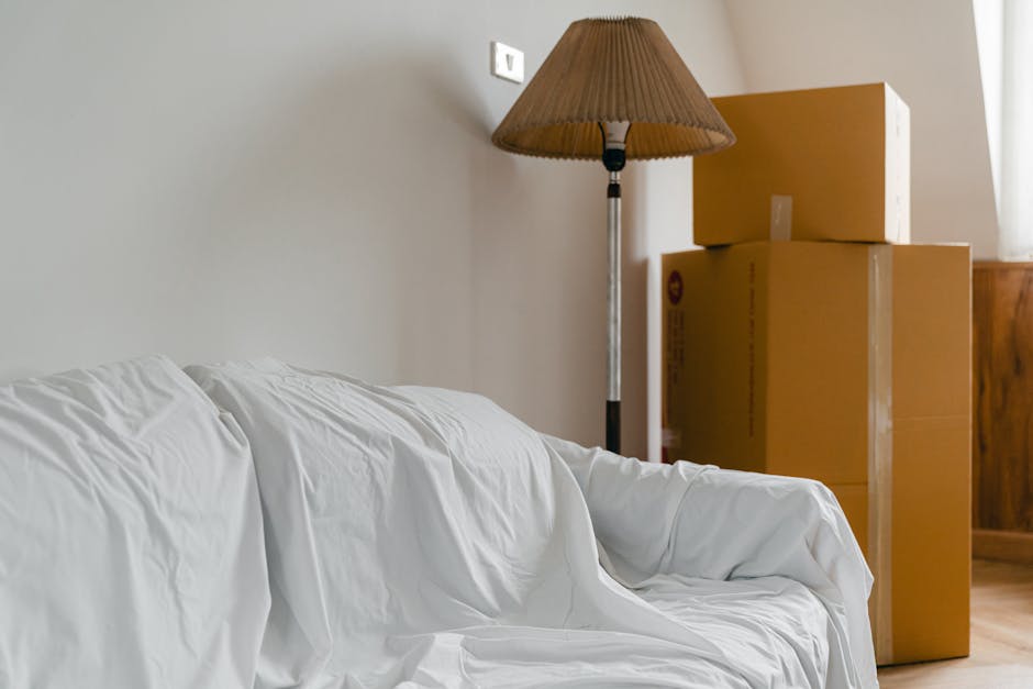 Inside a home during a furniture removal or home relocation process, there is a white sofa with a slipcover draped over it positioned on the left side of the image. In the background, three cardboard boxes of varying sizes are stacked against a beige wall near a wooden piece of furniture, likely a cabinet or sideboard. To the right of the boxes, a tall floor lamp with a brown fabric lampshade stands prominently, its metal stand extending from the floor. The room is well-lit by natural light coming from the right side, possibly through a window covered by a curtain or blind. The scene captures the stage of packing or unpacking in a property, with typical moving materials like cardboard boxes visible, supporting the context of professional removals by Man and Van Deptford, especially relevant to house removals or furniture transport services.