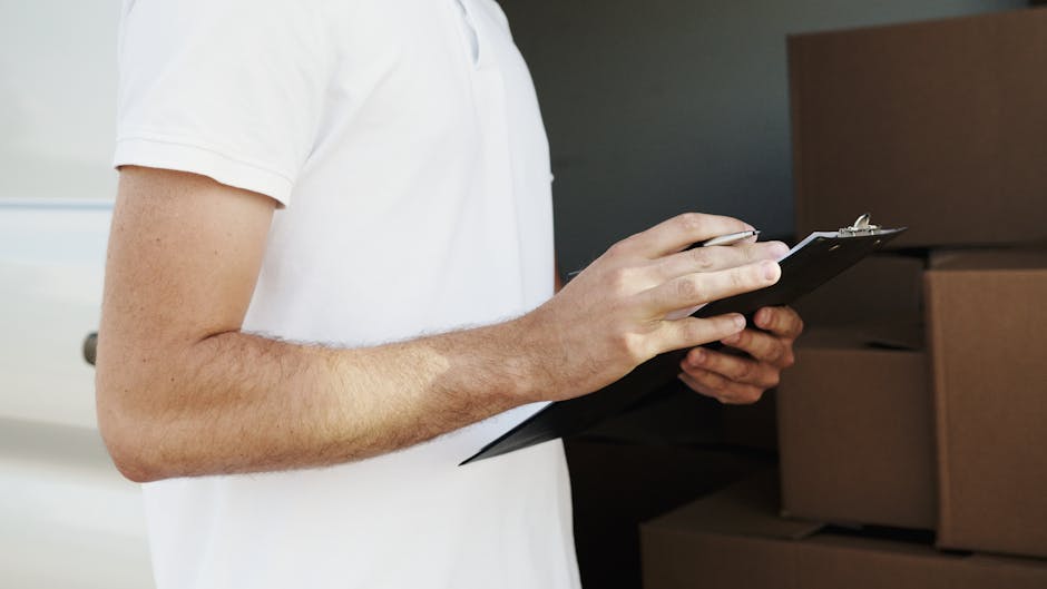 Inside a home during a furniture removal or home relocation process, there is a white sofa with a slipcover draped over it positioned on the left side of the image. In the background, three cardboard boxes of varying sizes are stacked against a beige wall near a wooden piece of furniture, likely a cabinet or sideboard. To the right of the boxes, a tall floor lamp with a brown fabric lampshade stands prominently, its metal stand extending from the floor. The room is well-lit by natural light coming from the right side, possibly through a window covered by a curtain or blind. The scene captures the stage of packing or unpacking in a property, with typical moving materials like cardboard boxes visible, supporting the context of professional removals by Man and Van Deptford, especially relevant to house removals or furniture transport services.