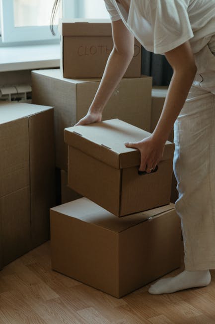 A person, dressed in light-colored clothing, is seen in an indoor setting, carefully handling a cardboard box during a house relocation process. The individual is bending slightly forward, gripping the box with both hands, with other stacked cardboard boxes surrounding them. One of the boxes has the word 'CLOTHES' handwritten on it, indicating its contents. The scene takes place next to a window with natural light illuminating the area, and the floor is made of wooden planks. This image reflects packing and handling activities typical of a professional moving service, such as those offered by Man and Van Deptford, as part of comprehensive home relocation and furniture transport solutions.