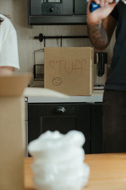 A person lying on the light wooden floor of a room, partially hidden under a stack of cardboard boxes during a home relocation process. The individual is wearing dark pants and socks, with their upper body obscured by the boxes. Several cardboard boxes of varying sizes, some with handles, are placed haphazardly around them, indicating packing and moving activity. The room features white walls, a large window allowing natural light to illuminate the space, and a closed white door with panel design. Behind the boxes, moving blankets and packing materials may be visible, and an area outside the window shows a glimpse of buildings or outdoor environment. The scene depicts the loading or packing stage of furniture transport, with careful handling of household items as part of a professional removals service, such as those offered by Man and Van Deptford.