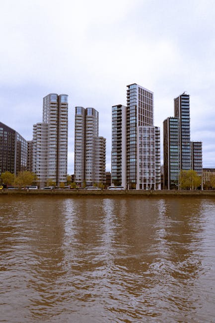 A row of modern high-rise residential buildings situated along the riverbank, with multiple floors and extensive glass window panels reflecting the overcast sky. In the foreground, the brown water of the river is visible, with gentle ripples on the surface. The buildings are positioned behind a narrow strip of pavement and sparse trees, indicating an urban riverside setting. The scene suggests a typical outdoor view during a house removal or furniture transport process, which could be part of a relocation project managed by Man and Van Deptford. Although no moving equipment or furniture are visible in this specific image, the urban landscape and riverside environment are relevant to logistics involving building moves or home relocation services in the Deptford area, as highlighted in the webpage about riverside building moves.