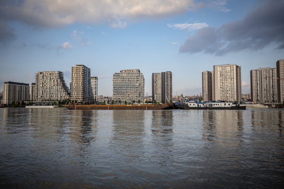 A panoramic view of a riverside urban landscape with modern high-rise apartment buildings lining the opposite bank of a wide river. The buildings feature contemporary architectural designs with varying heights, balconies, and large windows. The river in the foreground has a gentle ripple effect, reflecting the partly cloudy sky above with patches of blue and scattered clouds. Along the riverbank, there are some industrial structures and boats docked near the shoreline. The scene is illuminated by natural daylight, providing clear visibility of the buildings’ facades. This setting depicts the area around Deptford Creek, relevant for house removals and furniture transport as part of home relocation or building moves. Occasionally, Man and Van Deptford uses such environments during loading or unloading processes, where packaging materials like cardboard boxes and protective blankets may be positioned nearby for efficient packing and transportation.