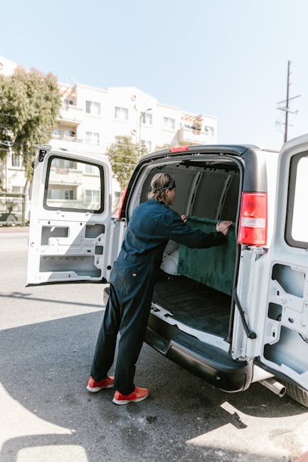 A person wearing dark clothing, including a blue jacket and black pants, is loading a large green cardboard box into the open cargo area of a white van with its side door wide open. The van is parked on a paved street during daylight, with residential buildings and trees visible in the background, indicating a home relocation or furniture transport scene. The individual is standing on the pavement, carefully positioning the box inside the vehicle, which is part of a professional removals process. Man and Van Deptford specializes in house removals, and this image illustrates the loading process involved in packing and moving household items using protective materials like cardboard, with equipment such as the van and possibly trolleys nearby for efficient logistics.