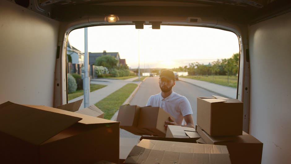 A man wearing a white polo shirt and sunglasses is seen loading cardboard boxes into the back of a van parked on a residential street during sunset. The open rear doors of the van reveal several packed cardboard cartons of various sizes, some with flaps open, ready for transport. The boxes are stacked inside the van, with additional boxes placed on the pavement beside the vehicle, indicating an ongoing home relocation process. In the background, suburban houses, a sidewalk, and a lamppost are visible, illuminated by warm, natural light. The scene captures the loading process typical of furniture transport and packing during a house move, with the man systematically placing boxes into the van, supported by the professional services of Man and Van Deptford. The image emphasizes the logistics involved in a professional removal and relocation service, highlighting careful packing and loading activities for a smooth move, with the van positioned for efficient access and cargo loading.