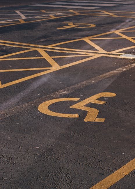 A close-up view of a parking lot surface showing yellow painted markings, including a large disabled parking bay with a wheelchair symbol in the foreground and additional lines and symbols for accessible parking spaces in the background. The asphalt surface appears slightly worn with visible texture and patches of grime. The scene is outdoors, illuminated by natural light. This image is relevant to house removals and furniture transport services, such as those provided by Man and Van Deptford, as it depicts parking areas often used for loading and unloading during property moves, and can relate to parking permit considerations for Lewisham Council and Deptford relocations.