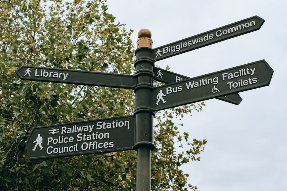 A black multi-directional signpost situated outdoors against a backdrop of leafy trees, with five separate signs pointing in different directions. The signs display white text and icons indicating nearby locations: 'Biggleswade Common', 'Library', 'Bus Waiting Facility', 'Toilets', 'Railway Station', 'Police Station', and 'Council Offices'. The 'Bus Waiting Facility' sign includes accessibility icons for wheelchair users and pedestrians. The scene appears to be in a public area, possibly near a transportation hub or community space, making it relevant for house removals and relocation services provided by Man and Van Deptford, as part of their furniture transport and packing logistics for home moves.