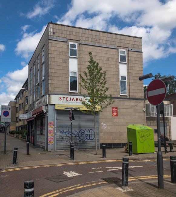 A three-storey building constructed with beige brick and large vertical windows on each level stands on a city street corner under a partly cloudy sky. The ground floor features a closed metal roll-up shutter with graffiti, and a sign above indicates a shop called 'Stejarul.' In front of the building, a small leafless tree is planted in the pavement, bordered by black bollards separating the pedestrian walkway from the road. To the right of the tree, a bright green waste or recycling bin is positioned next to a bench where a person is seated. The street area includes various traffic signs, such as a no-entry sign and a parking zone sign, and a black bicycle lane delineates the road. Visible in the background are additional buildings and parked vehicles, with the scene capturing a typical urban environment suitable for home relocation and furniture transport activities carried out by firms like Man and Van Deptford, particularly during the process of packing, loading, and moving household furniture and boxes in compliance with local regulations.