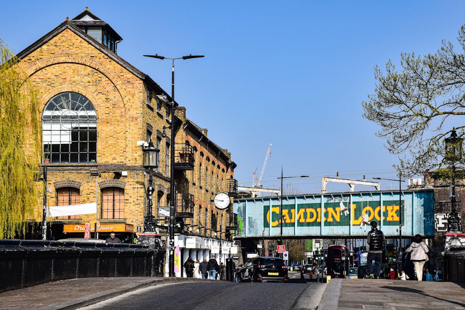 A street scene in Deptford showing a brick building with a large arched window and a clock mounted on a pole. In the foreground, there are pedestrians walking on the pavement, some carrying bags or wearing jackets, and a few are stopped near shopfronts. Several parked cars are visible along the street, with a black van near the curb. An overhead railway bridge with a green painted sign reading 'CALDIN LOCK' spans across the road, indicating a nearby train station or railway line. The sky is clear and blue, and trees with early spring foliage are visible on the right side. The scene captures typical urban activity related to home relocation and furniture transport, with the presence of vehicles and pedestrians involved in moving or logistics processes supported by companies such as Man and Van Deptford, as indicated on the site.