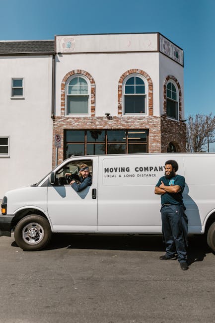 A white moving van parked in front of a two-storey residential building with a brick and white facade, featuring three arched windows on the upper floor and a large ground-floor window. Two men are present: one inside the driver’s seat, smiling and engaged in conversation, wearing a cap and jacket, and the other standing outside the van with arms crossed, dressed in dark clothing and smiling. The van displays the logo and text of a local moving company, and the scene takes place in a quiet, well-lit area during daytime, suggesting a home relocation or furniture transport process as part of packing and moving services offered by Man and Van Deptford, fitting with the context of house removals and logistics.