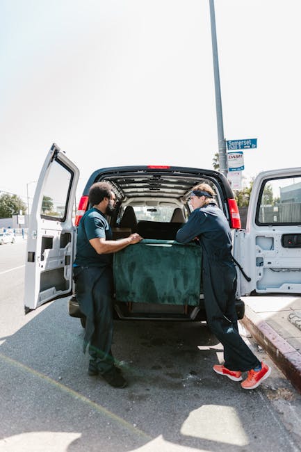 Two men engaged in a home relocation task are seen outside a residential property, unloading a large cardboard box from the back of a white delivery van parked on a brick driveway. One man, wearing a black vest and grey hoodie, is lifting the box while the other, dressed in a blue jacket and black vest, assists with guiding it. The van’s rear doors are open, revealing multiple packed cardboard boxes of various sizes inside, likely containing household items for a moving process. A hand truck with two wheels and a supporting frame is positioned nearby on the driveway, ready to facilitate further transfer of boxes and furniture. The scene is set in an outdoor environment with a red brick boundary wall, some shrubbery, and a house with a tiled roof in the background, under overcast weather conditions. This staged activity reflects the professional loading and packing steps involved in house removals, supported by companies like Man and Van Deptford, specializing in furniture transport and home relocation services designed for local estate moves.