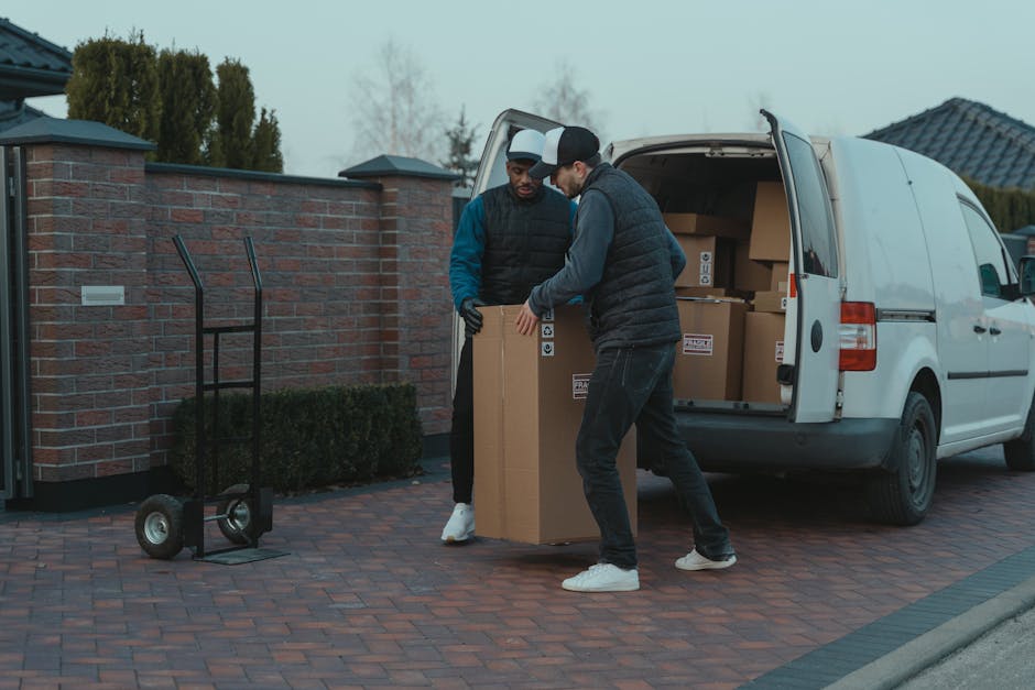Two men seated at the rear of a van during a home relocation, with cardboard boxes and packing materials visible in the background inside the vehicle. One man wears a beige beanie, a puffer vest over