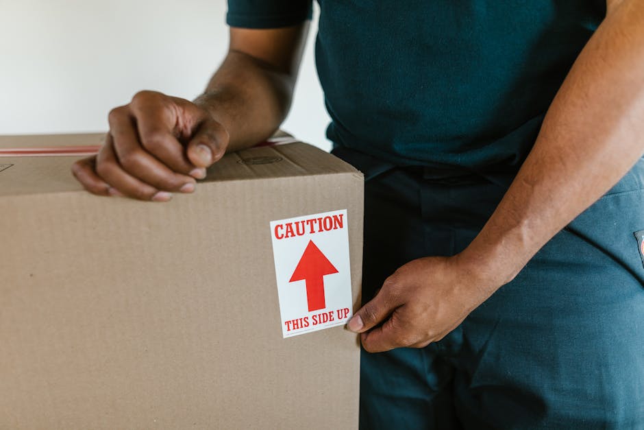 Close-up of a person wearing a dark green shirt and navy blue pants, sealing a cardboard moving box with packing tape. The box features a red and white caution label that reads 'CAUTION THIS SIDE UP' with an upward-pointing arrow. The individual is inside a well-lit room, standing near a doorway, preparing the box for transportation during a home relocation process. Behind the person, part of the room's plain white wall is visible, and the scene suggests an organized packing and loading process typical of house removals. The image captures the careful handling involved in furniture transport, packing, and loading activities carried out by Man and Van Deptford as part of their same-day removals service.