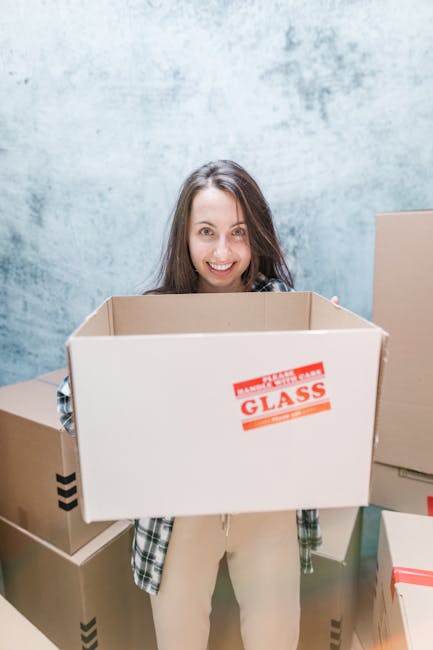 A young woman with long brown hair and a cheerful expression is standing indoors, holding an open cardboard box labeled 'GLASS' in red and black text. She is wearing a casual plaid shirt over a light-colored top. Surrounding her are several other cardboard boxes of various sizes, some sealed with packing tape and others open, placed on the floor and against a light grey textured wall. This scene depicts the process of home relocation, including packing and moving activities, with the woman preparing for furniture transport or a house removal. The environment is well-lit, suggesting a professional relocation setting as part of a same-day removal service in Deptford, coordinated by Man and Van Deptford.