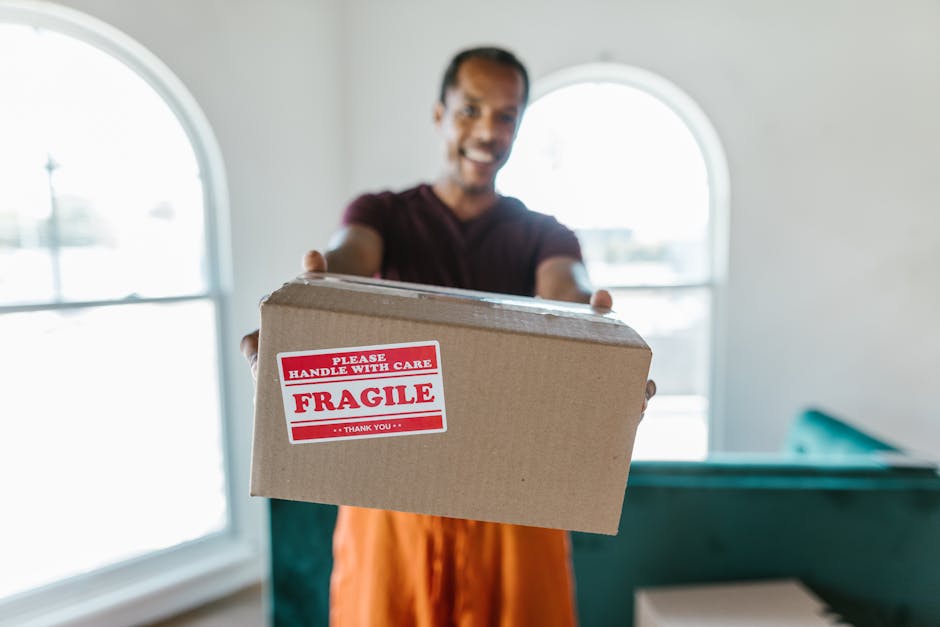 Close-up of a person wearing a dark green shirt and navy blue pants, sealing a cardboard moving box with packing tape. The box features a red and white caution label that reads 'CAUTION THIS SIDE UP' with an upward-pointing arrow. The individual is inside a well-lit room, standing near a doorway, preparing the box for transportation during a home relocation process. Behind the person, part of the room's plain white wall is visible, and the scene suggests an organized packing and loading process typical of house removals. The image captures the careful handling involved in furniture transport, packing, and loading activities carried out by Man and Van Deptford as part of their same-day removals service.
